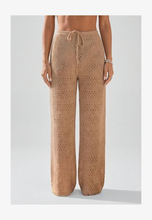 Pantalon large beige en maille crochet avec taille à cordon porté par une personne debout, raccourci au-dessus du ventre nu et des pieds.