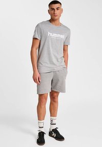 Grå bomullst-shirt med vit "hummel"-logotyp, kombinerad med matchande grå shorts. Accessoarer inkluderar mönstrade strumpor och svarta sneakers.