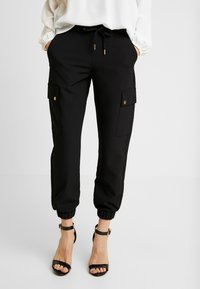 Mustat cargo-housut, joissa on joustavat hihnat ja kiristettävä vyötärö, kahdella sivutaskulla, joissa on kultaiset yksityiskohdat. Tasainen kankaan pinta.