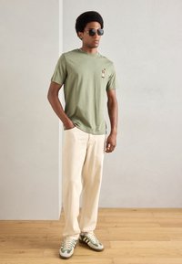 Olivengroen katoenen T-shirt met een borstafbeelding, gecombineerd met off-white spijkerbroek en sneakers met groene accenten, tegen een lichte achtergrond.