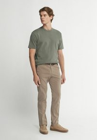 Grön bomullströja med kort ärm, i kombination medbeige chinos och bruna skor, står mot en neutral bakgrund.