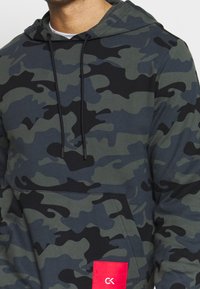 Homme portant un hoodie camouflage vert foncé et noir avec des cordons de serrage noirs et un patch rouge avec un logo CK blanc sur la poche avant.