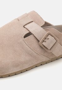 Beige mockasins i mocka med en texturerad yta, öppna sidor och en rektangulär spänne. Flexibel sula med en lågprofil design.