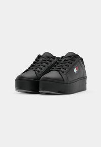 Zapatillas negras con material de cuero suave, punta redondeada, suela de plataforma gruesa, cordones en la parte frontal y un detalle de logo en azul, blanco y rojo.