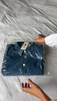 Denim-paita läpinäkyvässä muovipakkauksessa. Ominaisuuksiin kuuluu klassinen kaulus, edessä nappisuljenta ja etiketti, jossa lukee "Cotton Africa". Sininen väri.