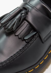 Mocasini din piele neagră, cu ciucuri, detalii împletite și cusături galbene de-a lungul tălpii. Textură netedă cu finisaj lucios.