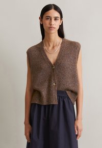 Brown knitted sleeveless vest with a loose fit, featuring a V-neck and two buttons at the front, paired with a dark blue layered skirt.