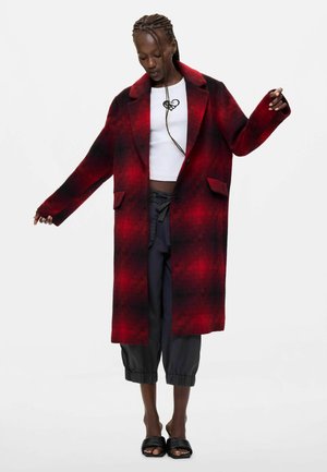 Donna che indossa un lungo cappotto a quadri rosso e nero, un crop top bianco con disegno di cuore nero, pantaloni neri corti e scarpe nere open toe.