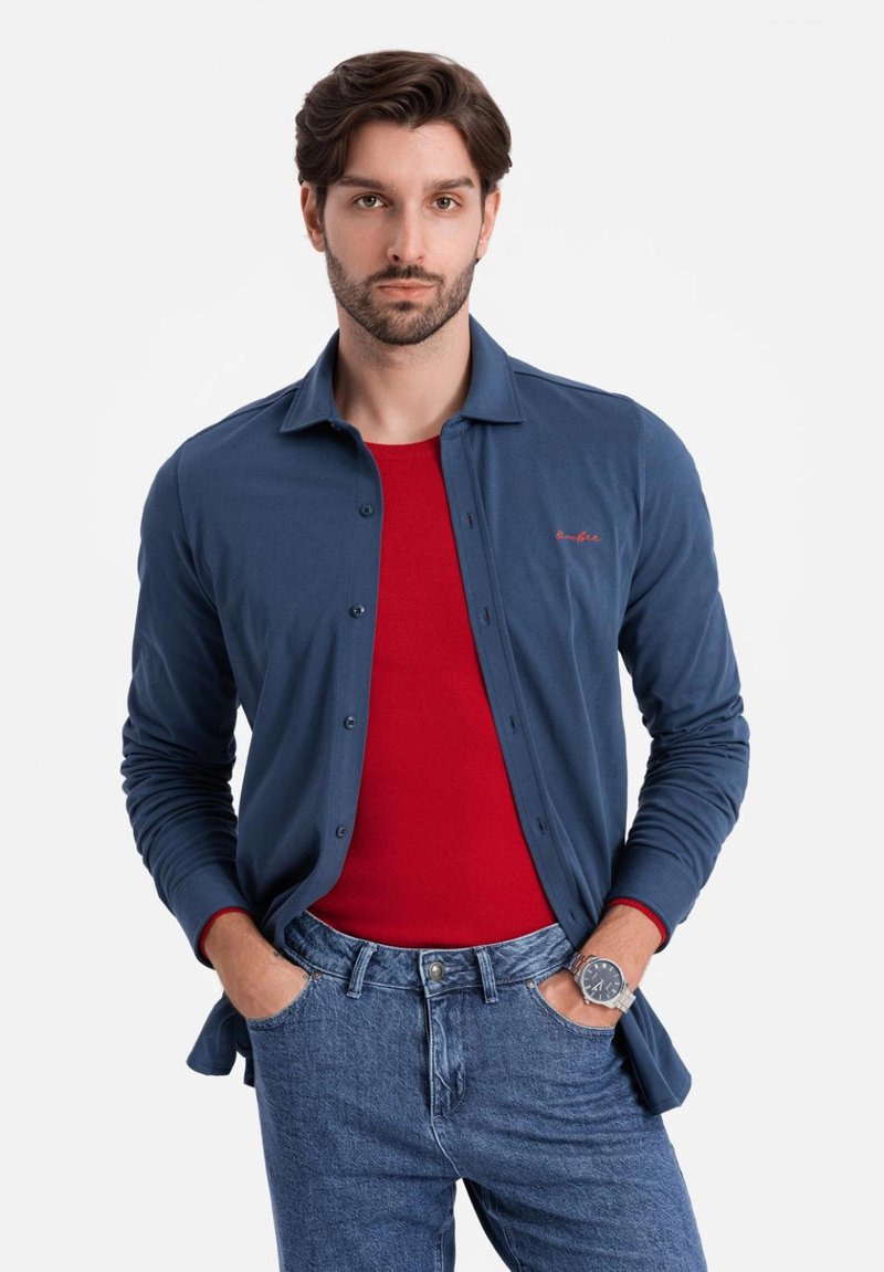 Camicia blu navy con bottoni sopra una maglietta rossa, abbinata a jeans blu. La camicia presenta una texture liscia e maniche lunghe, con un discreto logo.