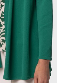 Grön, lätt långärmad tröja med en mjuk textur. Har en avslappnad passform och uppvikta ärmar, lagd över ett mönstrat plagg.