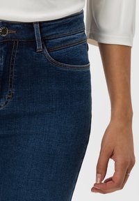 Mörkblå denimjeans med hög midja, kontrasterande söm detaljer, framfickor och en mjuk textur. Hand vilande på tyget.