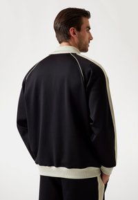 Sweatshirt noir avec un col crème, des poignets à côtes et des rayures sur les côtés. Texture lisse avec une coupe décontractée et un design minimaliste.