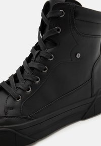 Černá vysoká teniska s texturovaným koženým svrškem, kovovými očky, plochými černými tkaničkami a silnou gumovou podrážkou.