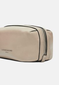 Beżowa skórzana kosmetyczka o gładkiej fakturze, z czarnymi zamkami błyskawicznymi i nadrukowanym brandingiem "LIEBESKIND BERLIN" z boku. Prostokątny kształt.