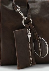 Donkerbruin leren tas met een bevestigde sleutelpouch met een zilveren rits en een sleutelcharme, plus een ronde metalen spiegelaccessoire.