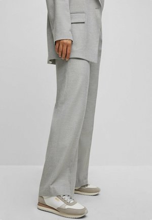 Persoon die lichtgrijze wijde broek, bijpassende blazer en beige-witte sneakers draagt, staand tegen een effen achtergrond.