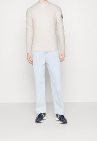 Vaaleanbeige, ribbineuloksinen neulepaita pitkillä hihoilla, yhdistetty vaaleansinisiin farkkuihin ja mustiin urheilukenkiin, jossa on pyöreä pääntie ja kädessä logo.