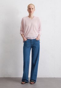 Helles rosa V-Ausschnitt-Sweatshirt mit langen Ärmeln, kombiniert mit hochgeschnittenen, dunkelblauen, weit geschnittenen Jeans und schwarzen Rutschsandalen.