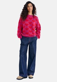 Chlupatý růžový svetr s červeným vzorem srdce, doplněný o široké tmavé džíny. Béžové boty s kontrastními tkaničkami a černými podrážkami.