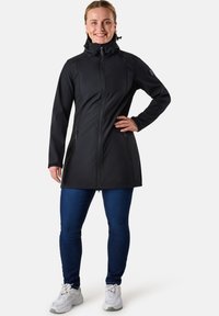 Zwarte softshell jas met hoge kraag, rits aan de voorkant en zijzakken; gecombineerd met blauwe jeans en witte sneakers.