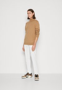 Maglione marrone a collo alto, pantaloni bianchi e sneakers. Il maglione ha polsini e orlo a coste; le sneakers presentano una combinazione di colori marrone e bianco.