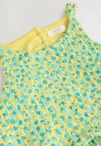 Gul utan ärmar topp med blått blommönster, ribbad textur och knappstängning på baksidan. Etiketten läser "NEXT, TILLVERKAD I SRI LANKA."