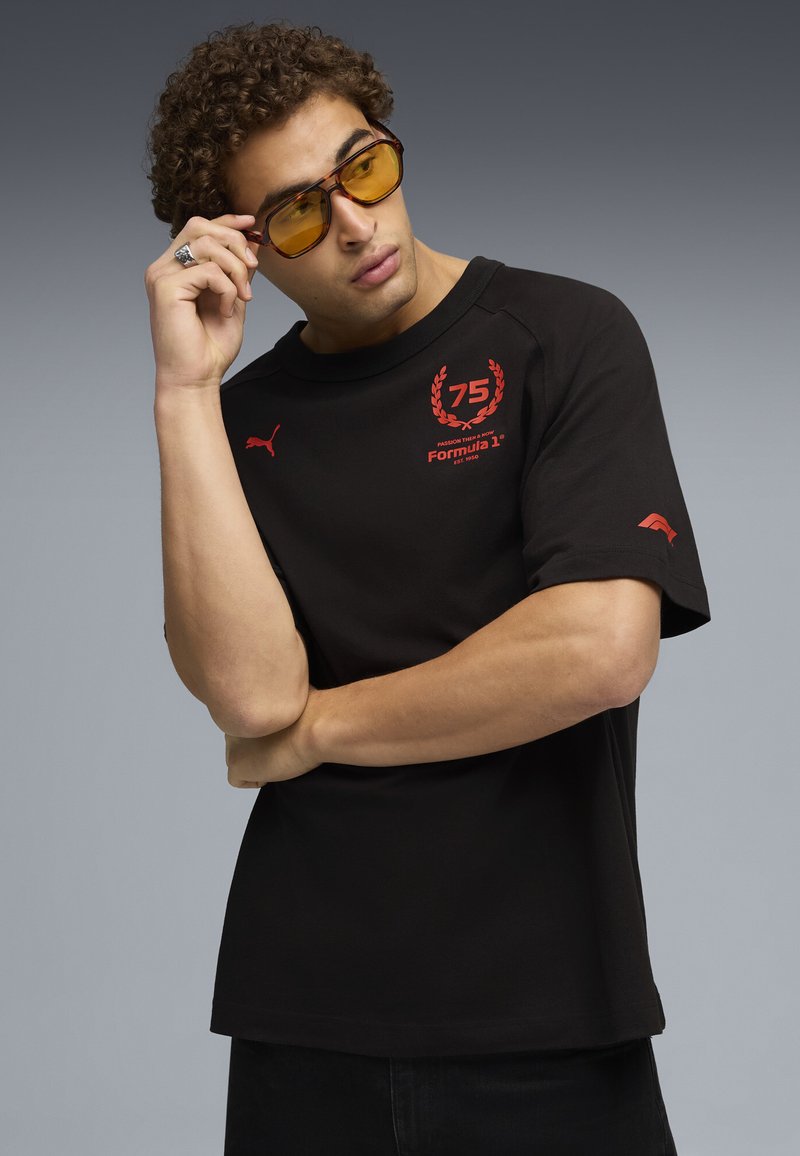 Musta lyhythihainen t-paita punaisella logolla ja yksityiskohdilla. Pyöreä pääntie, väljä mitoitus ja puuvillamateriaali. Yhdistetty keltaisiin sävyihin vaaleisiin aurinkolaseihin.