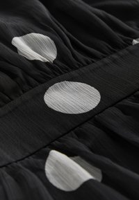Tessuto nero con motivi a pois bianchi circolari e texture arricciata lungo la cintura. Materiale leggero e fluido.