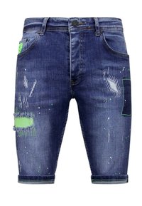 Donkerblauwe denimshorts met versleten details en groene accenten. Kenmerken zijn onder andere zakken, een knoopsluiting en rafelige hemden met verfspetters.