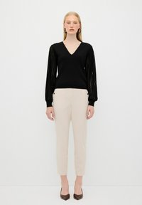 Schwarzer V-Ausschnitt-Pullover mit strukturierten, geplüschten langen Ärmeln, kombiniert mit cremefarbenen, schmalen Hosen und braunen spitzen Pumps. Einfaches und modernes Design.