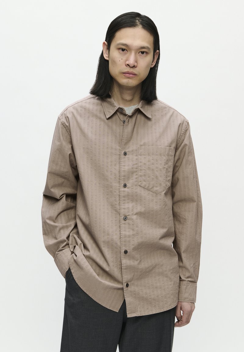 Camisa de botones a rayas marrones con cuello, un bolsillo en el pecho y camiseta gris debajo. El modelo lleva unos pantalones oscuros y tiene el cabello largo y lacio.