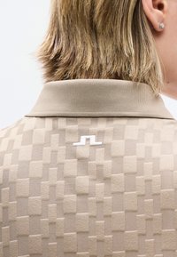 Μπεζ πικέ πόλο με ανάγλυφο γεωμετρικό σχέδιο, λευκό λογότυπο στο πάνω μέρος της πλάτης, φορεμένο από άτομο με κοντά ξανθά μαλλιά και σκουλαρίκι καρφίτσα.