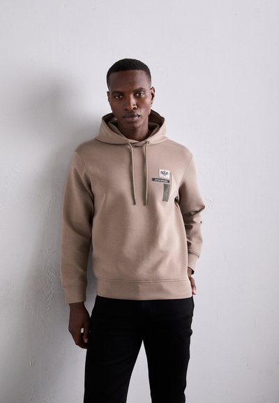 Hanorac bej cu glugă, având un buzunar în față, decorat cu un patch logo și etichete decorative; combinat cu pantaloni negri, cu o textură de țesătură netedă.