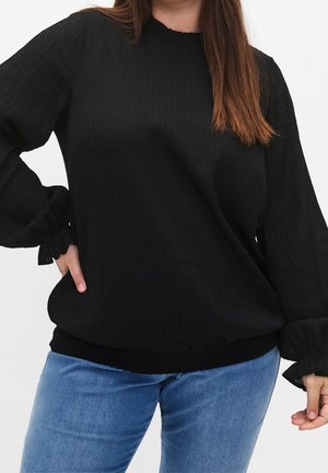 Femme portant un chemisier noir texturé à manches longues avec poignets et ourlet élastiques, associé à un jean bleu clair sur un fond uni.