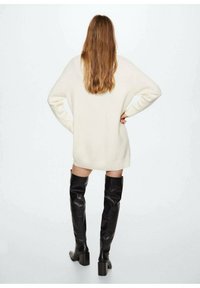 Oversized crèmekleurige gebreide trui met ribgebreide zoom; gecombineerd met kniehoge zwarte leren laarzen met chunky hakken; eenvoudige achtergrond.