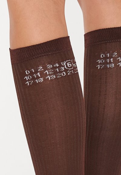 Pruunid põlvikud kuni põlvedeni, millel on soonikkoes tekstuur ja ülemises ribas on prinditud valged numbrid ja sümbolid.