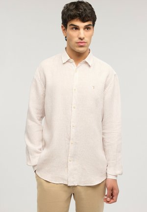 Jeune homme aux cheveux bouclés foncés portant une chemise à rayures beige clair boutonnée et un pantalon beige, debout devant un fond uni.