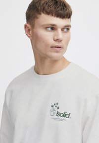 Crewneck sweatshirt i lys grå stoff har et grønt plantegrafikk med tekst på venstre brystområde. Jevn tekstur, avslappet passform.