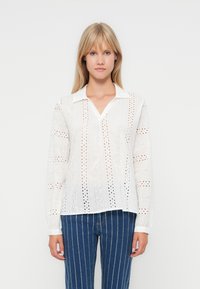 White long-sleeve blouse featuring broderie anglaise design, with a pointed collar and V-neckline. Textured fabric and relaxed fit.