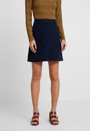 Kvinde iført en stribet brun langærmet top, navy knælængde nederdel med lommer og brune hælede åbne sandaler.