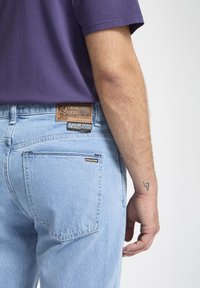 Jean en denim bleu clair avec une pièce en cuir beige présentant un motif embossé et des étiquettes. La poche arrière comprend une petite étiquette noire "VOLCOM".
