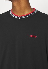 Schwarzes T-Shirt mit Rundhalsausschnitt, das einen kragen aus rot und schwarz gemustertem Strick hat. Das Levi's-Logo ist in Rot auf der Brust gestickt.