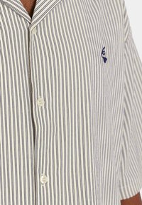 Camisa de manga corta a rayas en crema y azul marino. Presenta un cuello con muesca, cierre de botones y un logotipo bordado en el lado izquierdo del pecho. Tejido suave.
