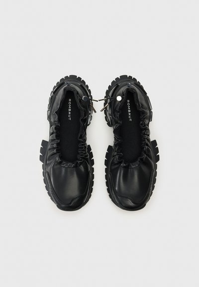 Black slip-on shoes with a gathered upper, textured rubber soles, elastic accents, and minimal branding in white on the insole.