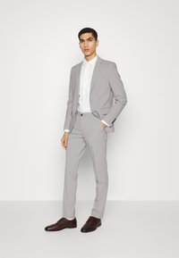 Completo grigio chiaro con una vestibilità su misura, tessuto strutturato e rever a lancia; abbinato a una camicia bianca con bottoni e scarpe in pelle marrone.