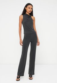 Pelēks bez piedurknēm jumpsuits ar vērpšanas apkakles detaļu, platām biksēm un gludu, mīkstu tekstūru, kombinēts ar melniem sandaliem.