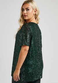 Top verde con paillettes, maniche corte, scollo rotondo e design a vestibilità ampia. La superficie scintillante riveste il capo, riflettendo la luce.