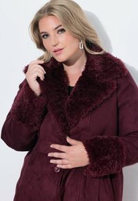 Bordeauxfärgad kappa med krage och manschetter i konstpäls, mjuk känsla, bälte i midjan och knappstängning i ett slätt, mockaliknande material.