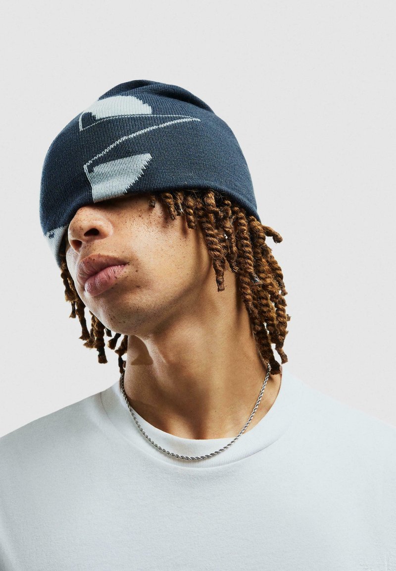Beanie a maglia in blu navy con motivo astratto bianco, texture morbida, design aderente e forma leggermente cadente, indossato basso sul viso.