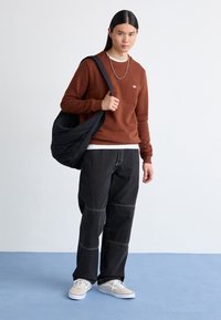 Brown sweatshirt with a small logo, black baggy pants with white stitching, and beige sneakers. Model holds a black bag.